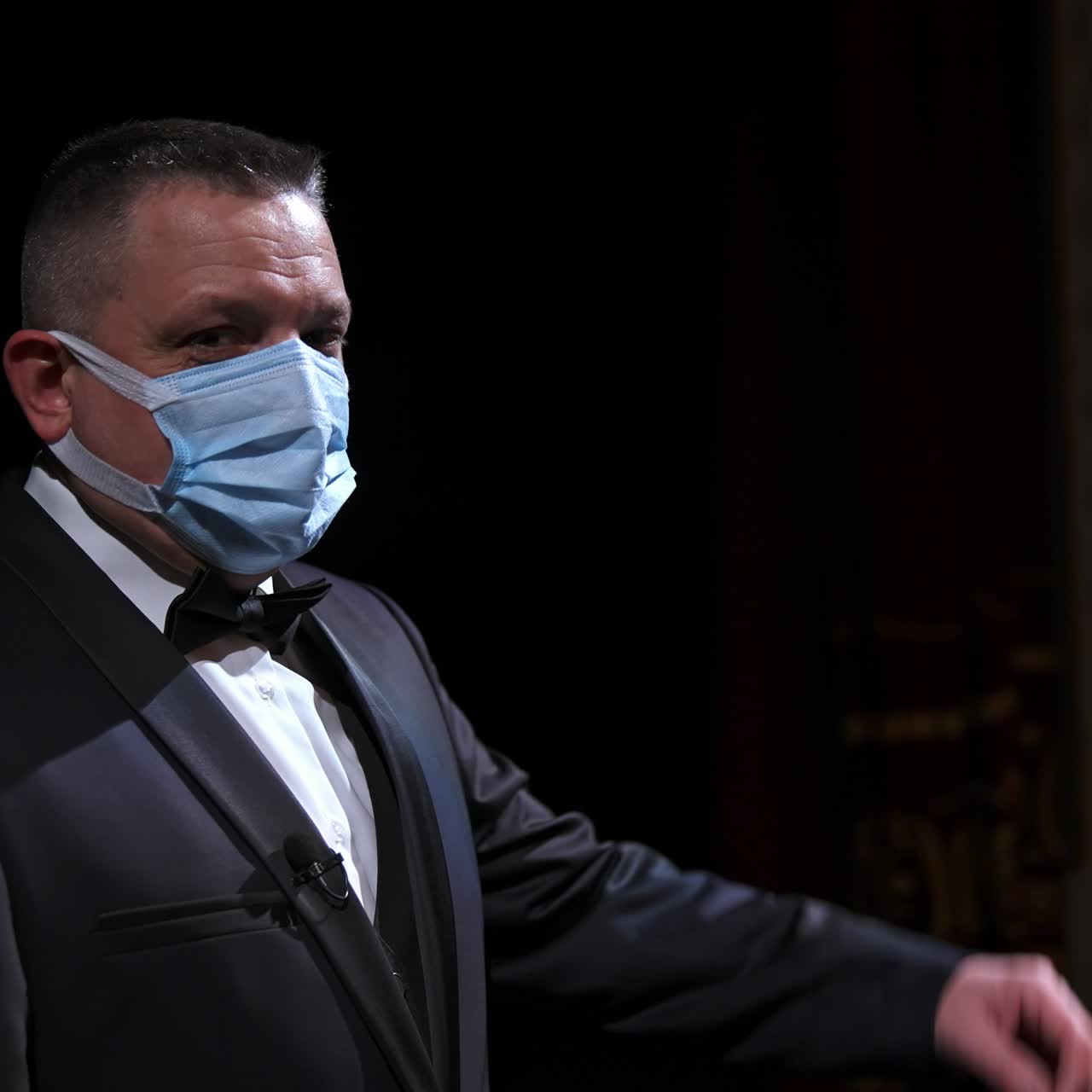 Ator de teatro fala no palco usando máscara médica. Apresentador de terno parado em frente a um salão de teatro vazio. Conceito de pandemia