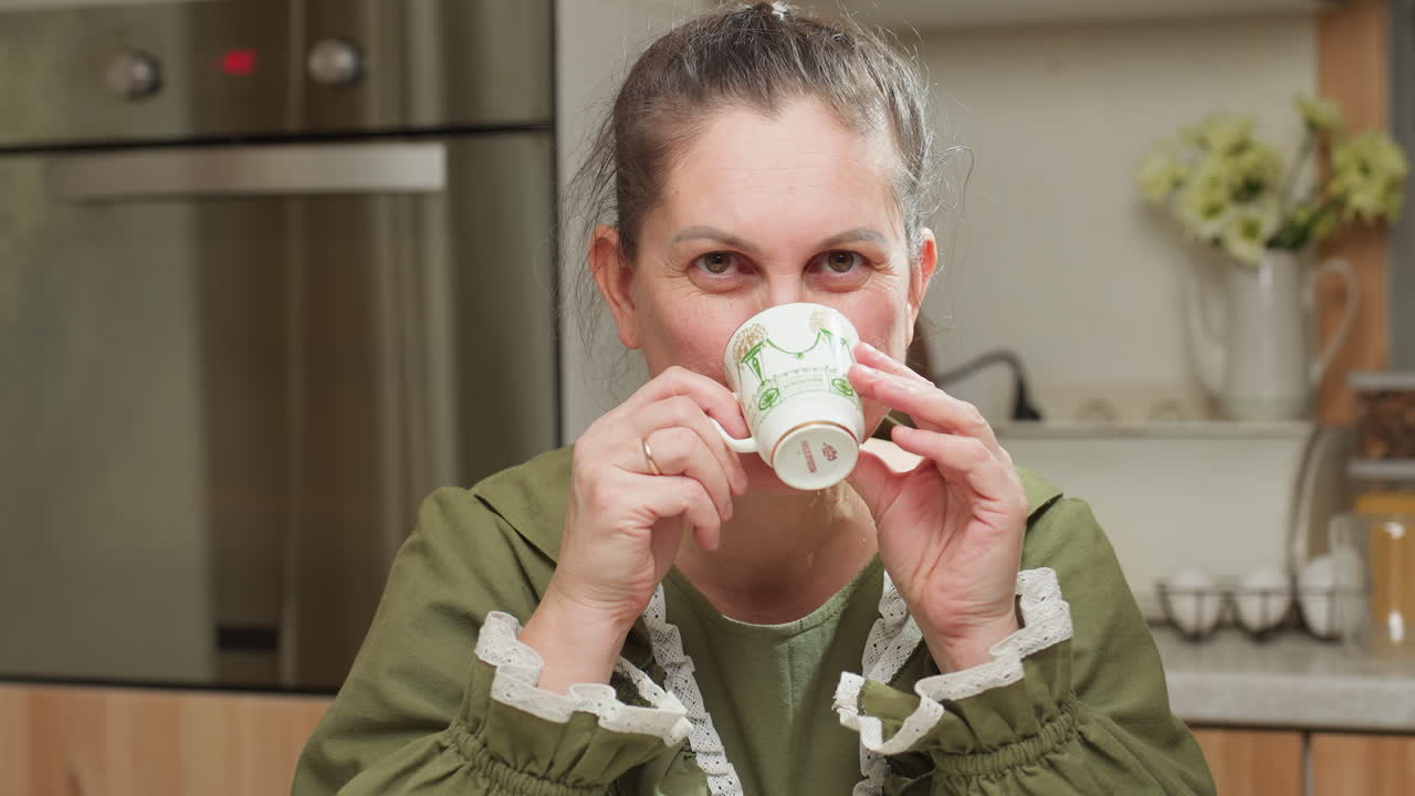 Young girl in green vintage-style dress enjoying hot coffee while gazing directly at camera with warm expression in cozy kitchen ambiance, creating inviting and comforting breakfast moment
