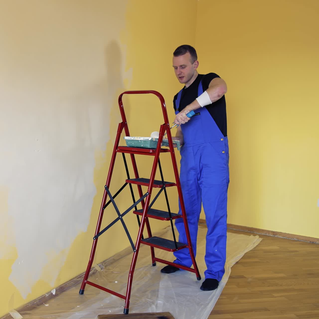 Young man in overalls renovating room in the house. Painter worker using a paint roller doing makeover of a room. Room decoration with new color