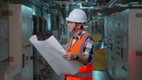 side view of asian male engineer with safety helmet looking at blueprint in his hands and looking around while standing in engine control room, work of electrical generators