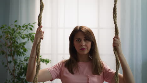 serious brunette woman sits on swing holding ropes at home