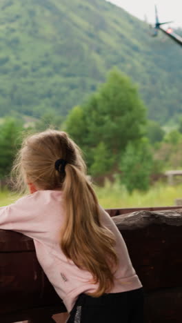 curious little girl looks at flying up helicopter leaning on large wooden log railing on terrace of rural cafe on cloudy summer day slow motion