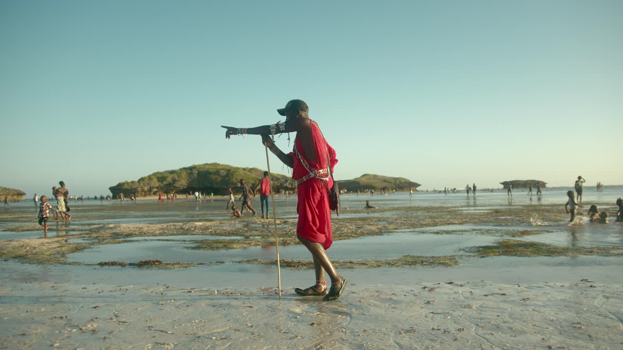 guerrero masai con ropa tradicional roja parado en la playa sosteniendo un poste de madera y apuntando en dirección a watamu, kenia