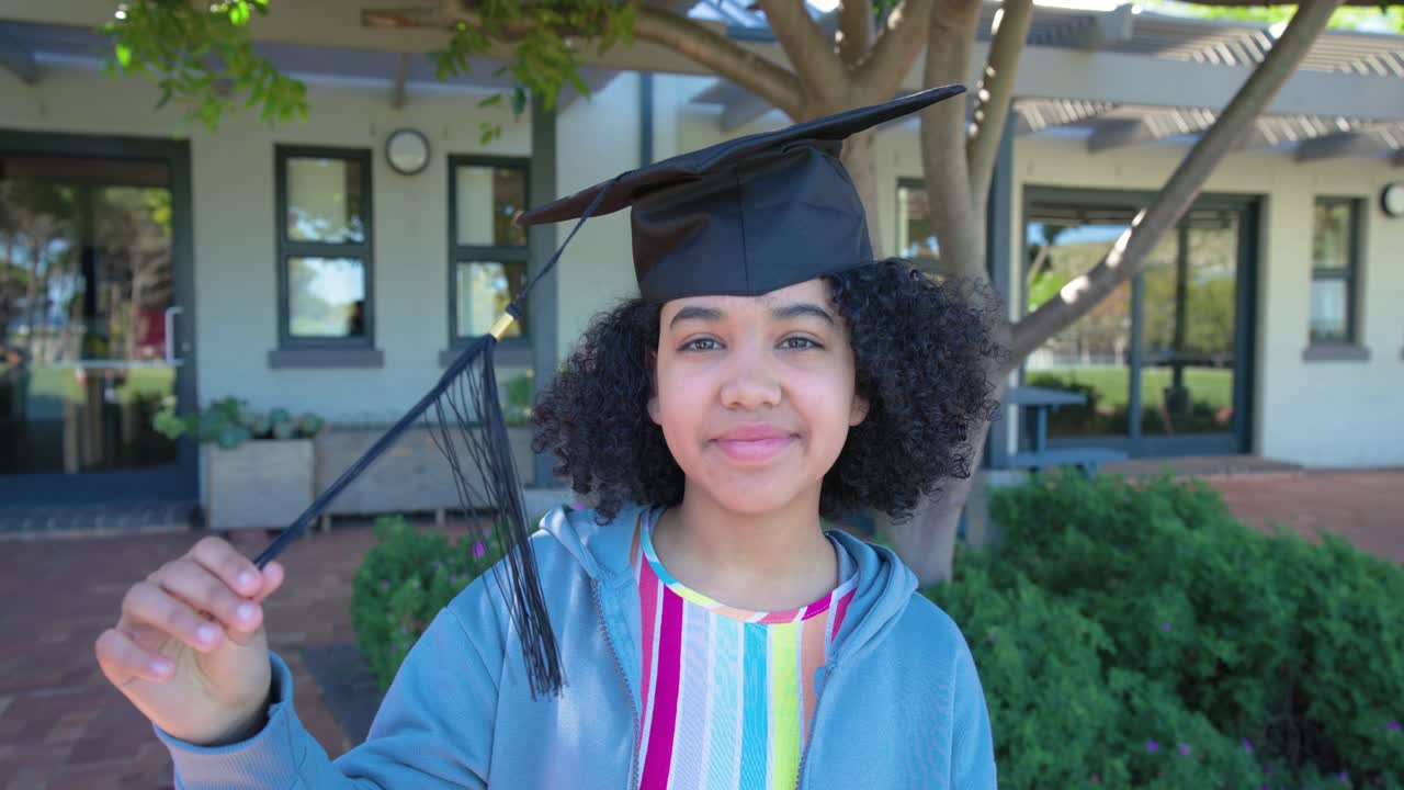 Adolescente afro-americana perto de um prédio, usando boné, inclinando a cabeça, ajustando a borla e balançando-a.