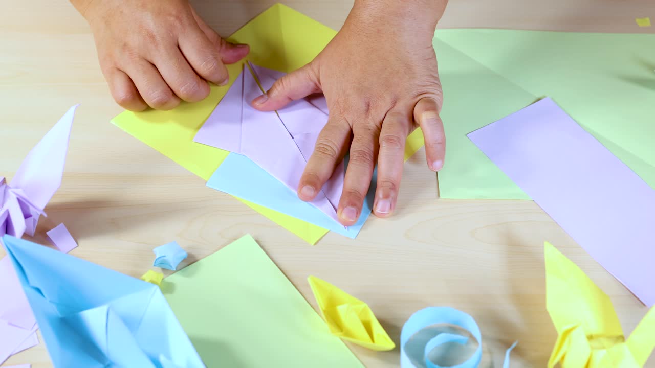 Hands fold pastel paper into origami shapes on a table, bright lighting, overhead camera view