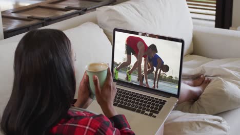 Composite-of-woman-sitting-at-home-holding-coffee-watching-athletics-running-event-on-laptop