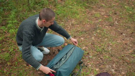 overhead angle shows young man in green knitted sweater crouching on forest floor carefully opening tent bag in woodland, surrounded by grass and leaves, preparing for outdoor camping activity