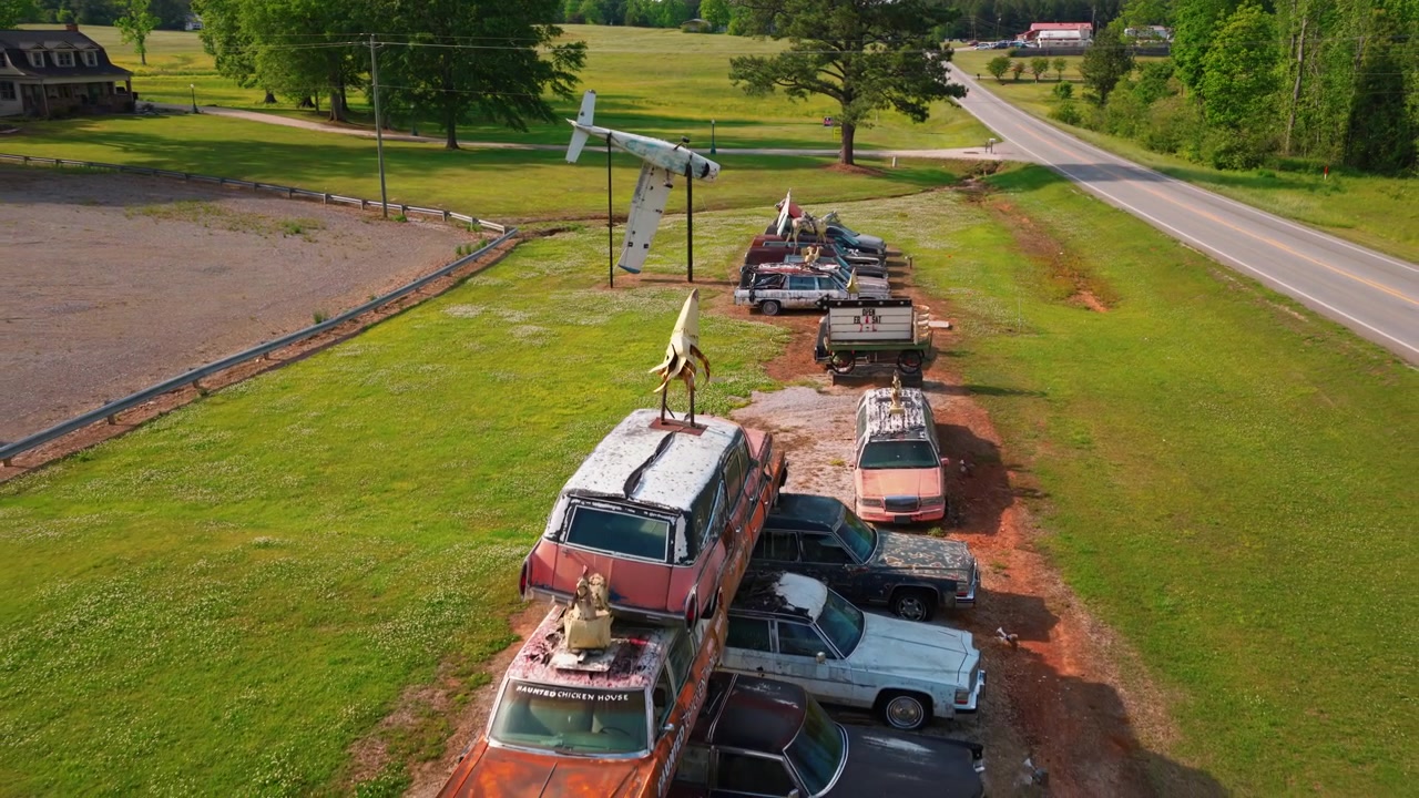 A surreal aerial POV shot of a haunted roadside attraction in Alabama featuring a stack of rusted vintage cars and a crashed airplane in a grassy field.
