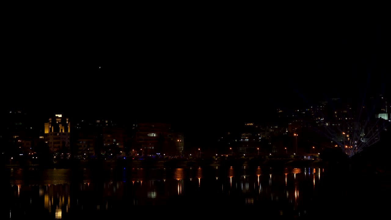 Fari laser che illuminano il cielo notturno sopra la città vicino alla superficie d'acqua riflettente a Tirana