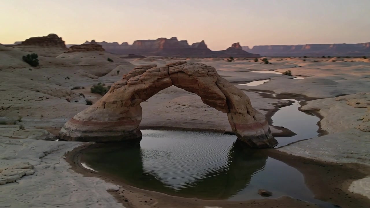 La telecamera si allontana e si sposta a destra mentre la luce si ammorbidisce, rivelando un arco di arenaria sopra una pozza d'acqua poco profonda.