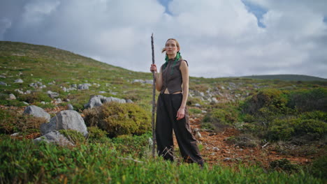 chica de senderismo posando colina de montaña con palo. aventurero de moda de pie verde