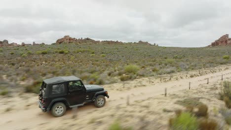 drone flies sideways and quickly tracks a black off-road vehicle on the road in cederberg wilderness area in south africa - rocky mountains can be seen in the background