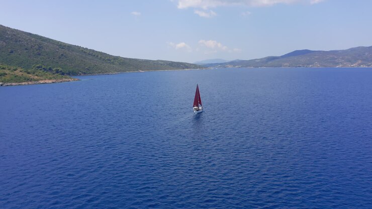Aereo: Ripresa lenta con drone di una barca a vela con vele rosse che naviga al largo in una limpida giornata estiva