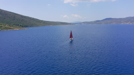 Aerial:-Slow-panning-drone-shot-of-a-sailboat-with-red-sails-that-is-sailing-offshore-on-a-clear-summer-day