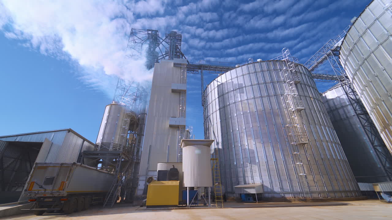 Contemporary factory for agribusiness. Steel elevators for grain processing outdoors. White smoke filling the sky from industry. View from below.