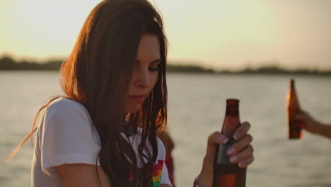 a female student with beautiful gray eyes is dancing on the open air party with her friends and beer. she smiles and touchs her long dark hair and enjoys summertime on the river coast at sunset.