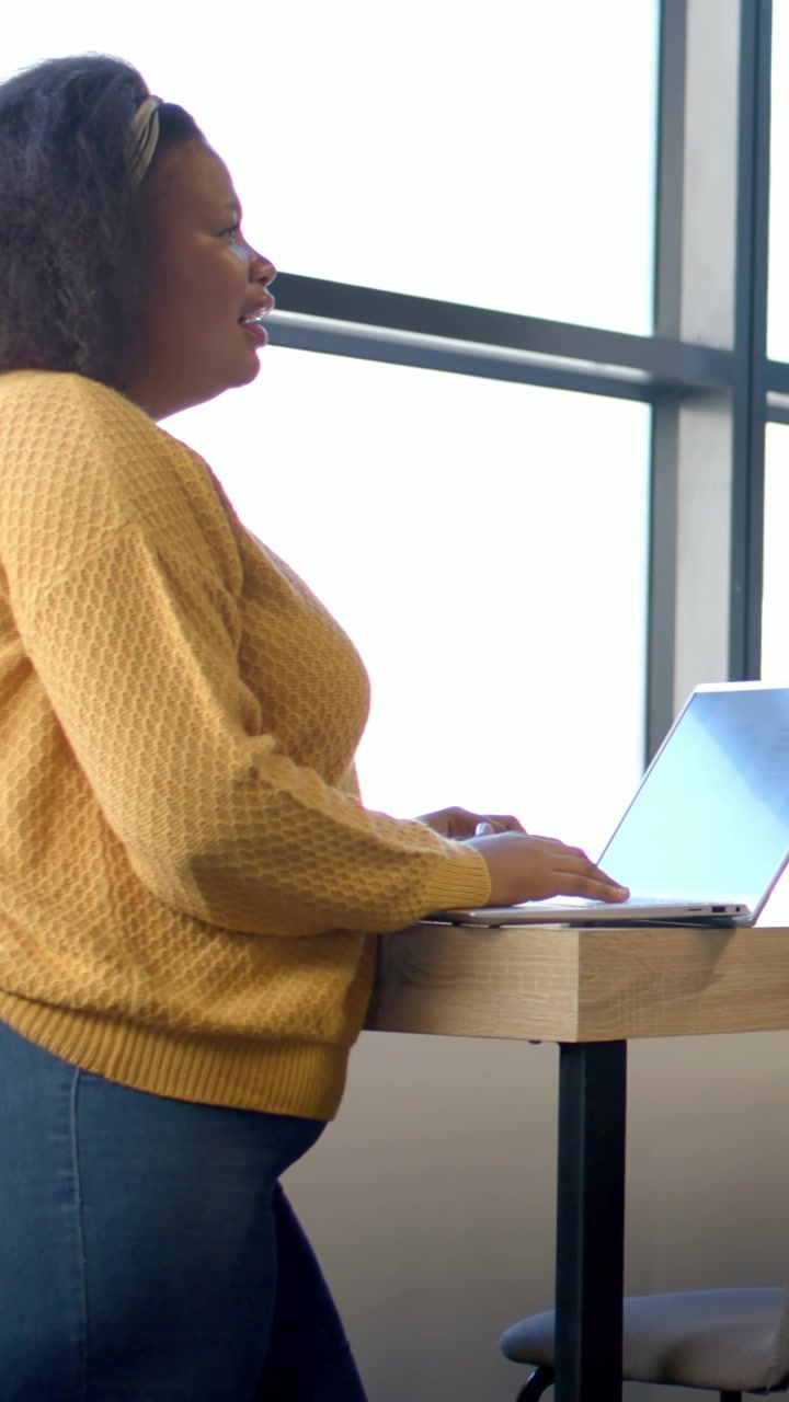 Video verticale: Inquadratura che parte dal laptop, lei si china, digita e parla al telefono vicino alla finestra, indossando un top color senape.