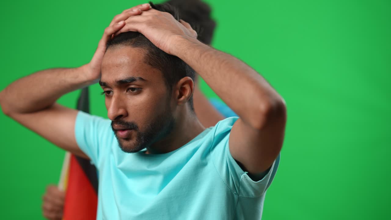 Focused sport fan looking away throwing away ball holding head in hands. Portrait of young Middle Eastern man watching match with friend waving German flag at background on green screen.