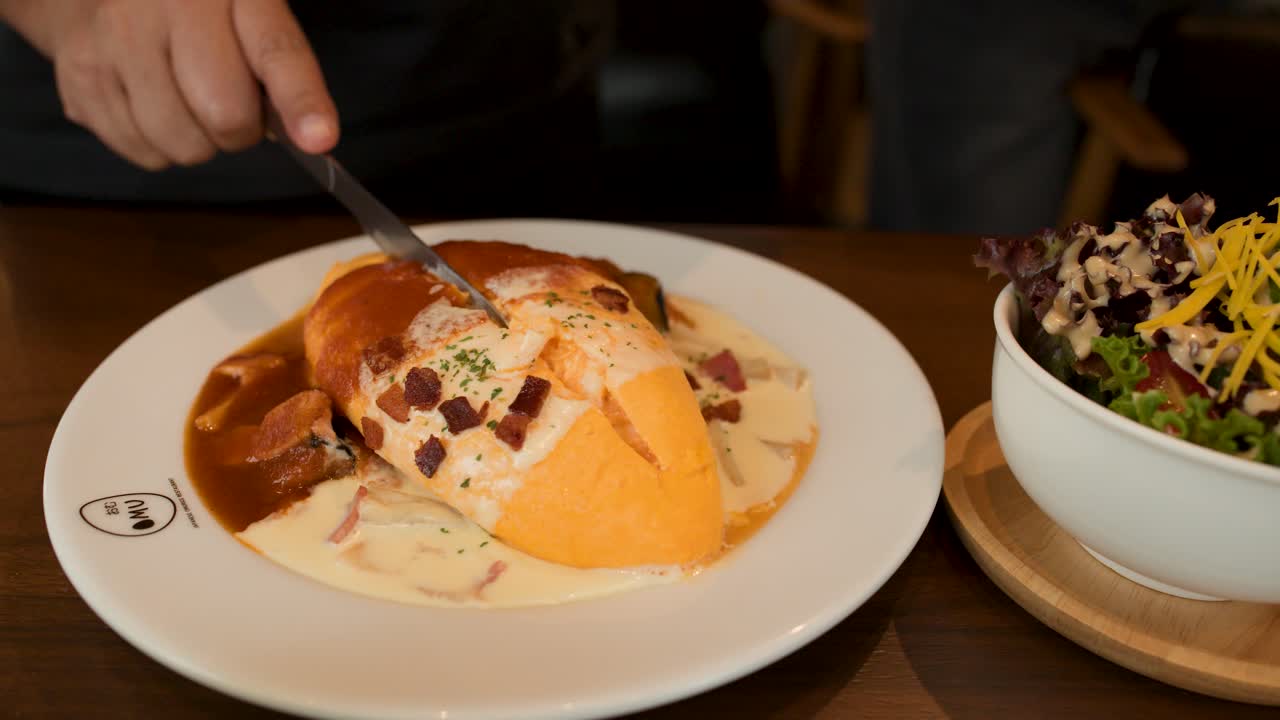 Hand slices omelette rice with creamy sauce in restaurant, warm lighting, close-up view