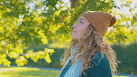mujer joven tranquila con sombrero y bufanda relajándose y respirando profundamente en el parque de otoño