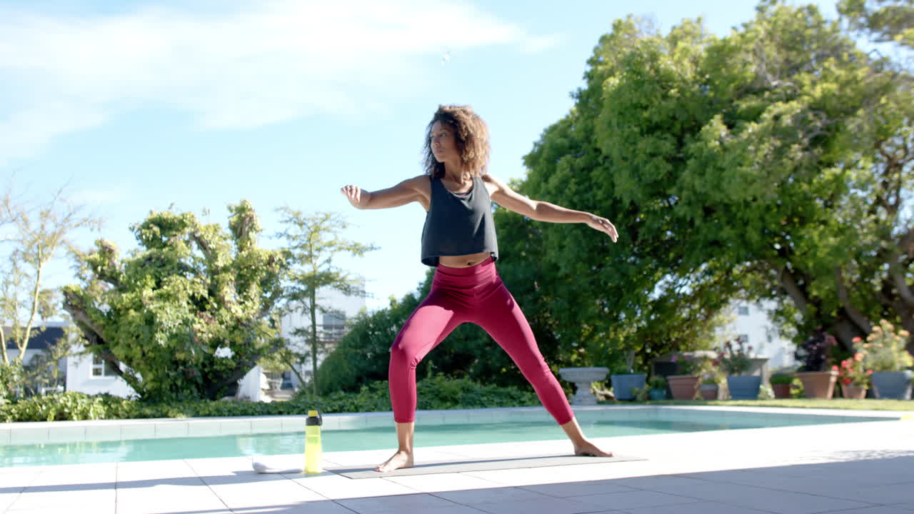 feliz mujer biracial practicando yoga de pie junto a la piscina en el jardín soleado, espacio de copia, cámara lenta
