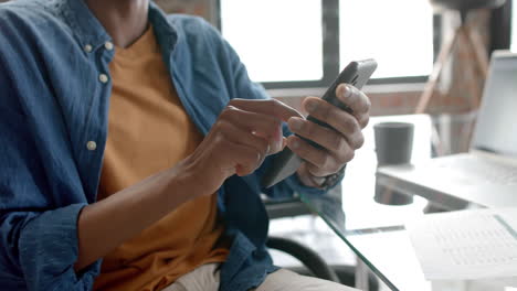 midsection of african american casual businessman sitting using smartphone at home, slow motion