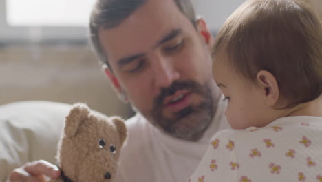 happy couple lying in bed on sunday morning and playing with their cute baby girl using a teddy bear