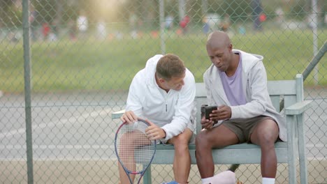 happy diverse male friends looking at smartphone on bench at tennis court, slow motion