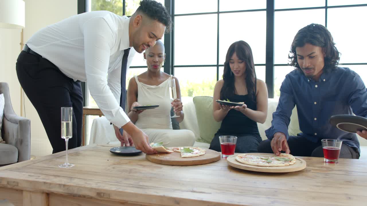 Diverse friends sharing pizza in living room, standing man white shirt placing slices for guests