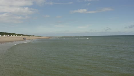 Toma-Aérea-Moviéndose-Sobre-Las-Olas-En-El-Mar-A-Alta-Velocidad,-A-Lo-Largo-De-Una-Playa-Donde-La-Gente-Camina-En-Un-Hermoso-Día-Soleado