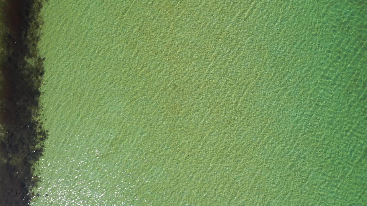 Aerial shot looking sown at the shallow turquoise sea, starting low and rising up on a bright summers day
