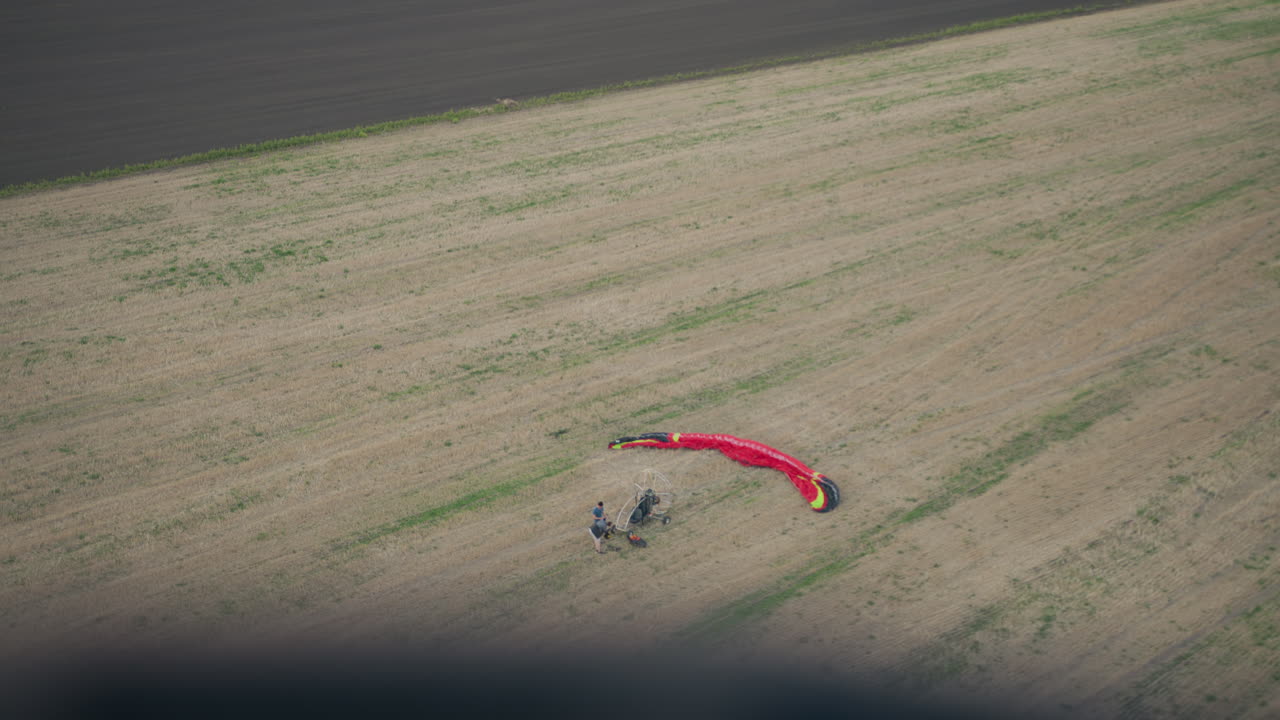 Top down view of people preparing vibrant red paraglider canopy on open field next to motorized fan cart, hands arranging lines and equipment on grassy terrain under gentle evening light