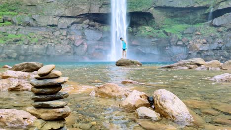 Un-Hombre-Aislado-Sentado-En-Una-Roca-Con-Una-Cascada-Que-Cae-De-La-Montaña-Con-Un-Montón-De-Piedras-En-El-Día-Se-Toma-En-Meghalaya