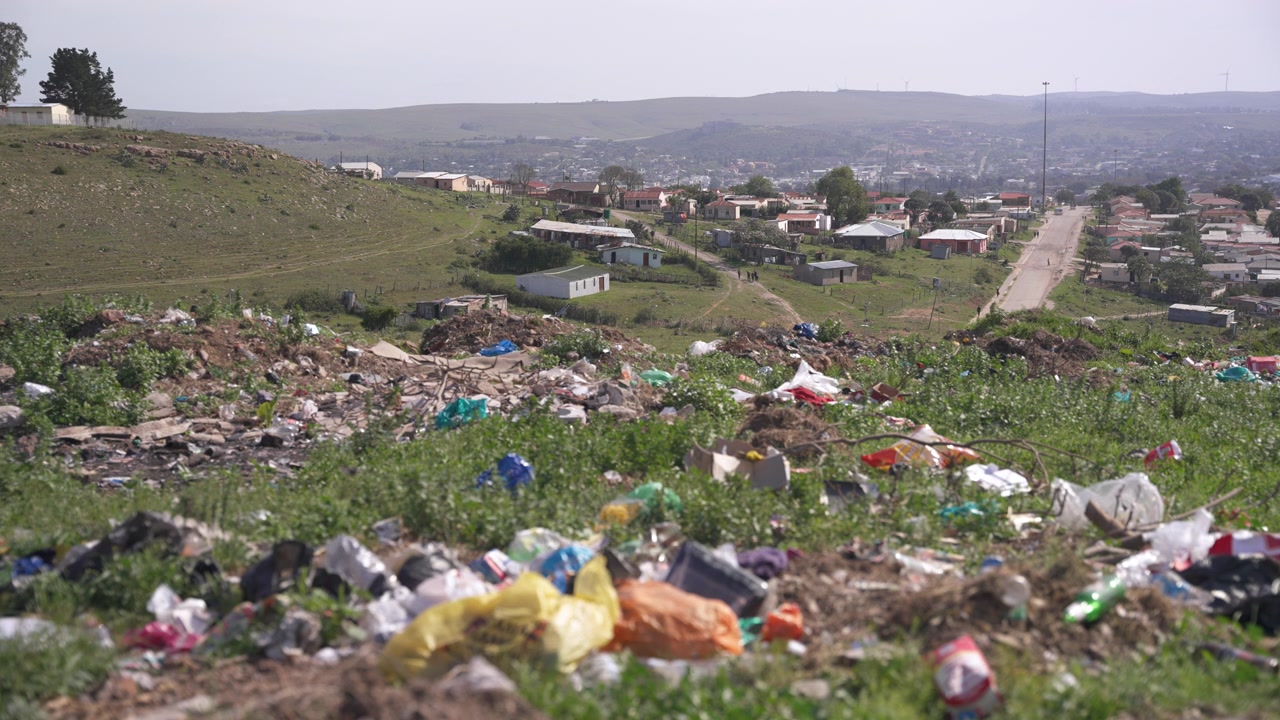 Plastic packaging and mixed rubbish cover a grassy hillside while modest homes and a long residential street stretch into the distance.