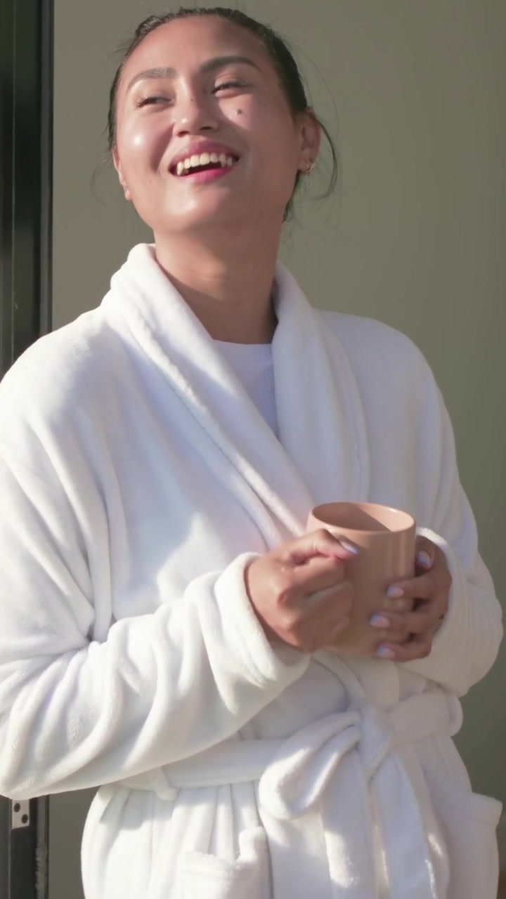 Vertical video: Sunlight waking woman in bathrobe, opening eyes and sipping tan mug at sliding door
