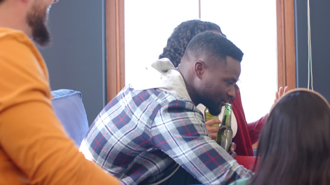 Happy diverse male and female friends talking at home, drinking beers and eating popcorn
