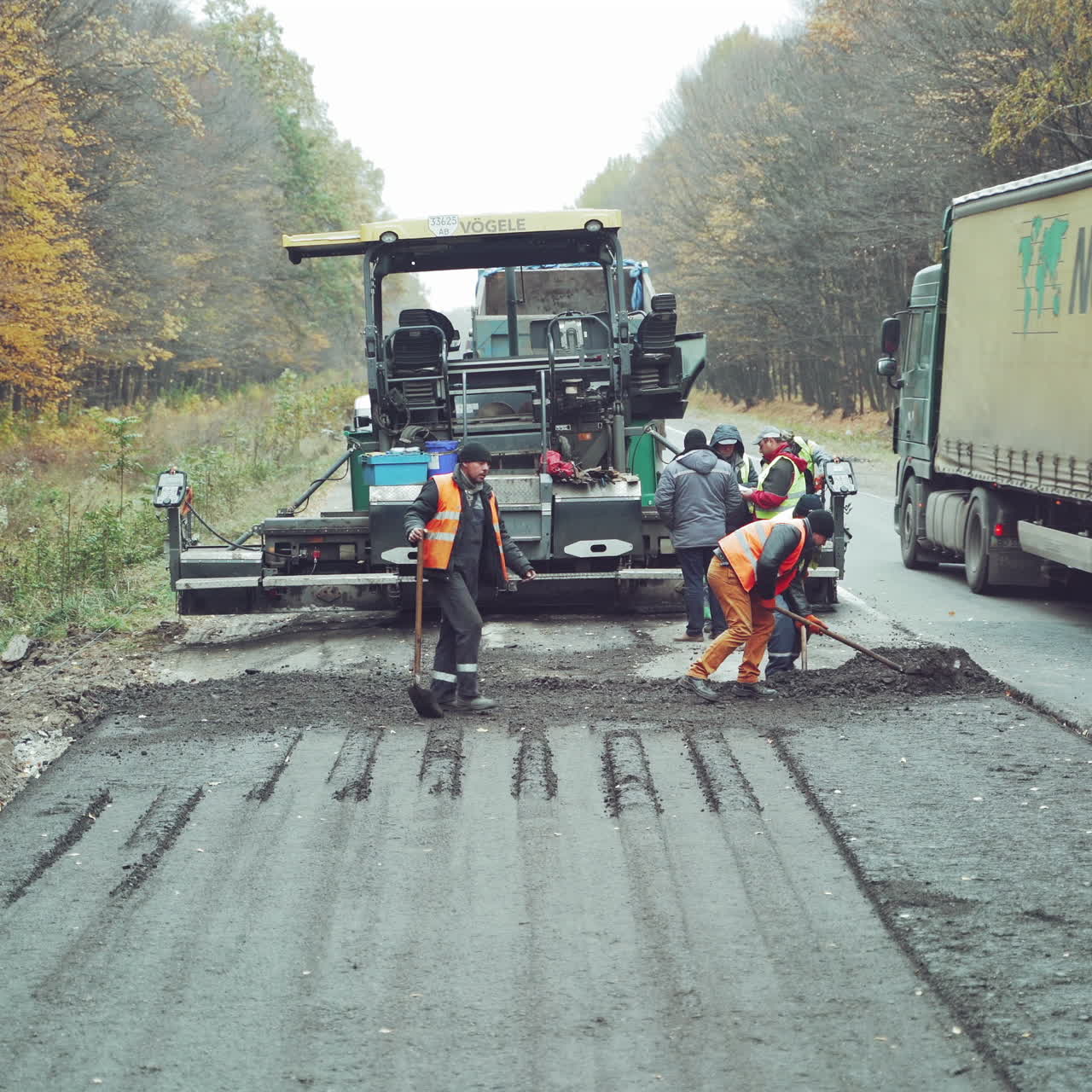 Team of workers put the hot asphalt on a half of the road on the background of a big steamroller. Road construction workers with shovels in protective uniform and the truck going by in autumn.