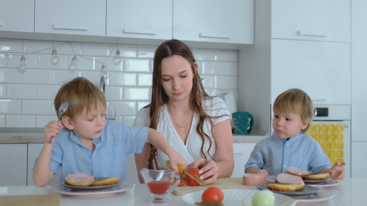 una familia feliz es una joven madre hermosa con un vestido blanco con dos hijos con camisas azules preparando una cocina blanca juntos cortando verduras y creando berger saludable para los niños.