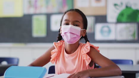 portrait of mixed race schoolgirl wearing face mask, sitting in classroom looking at camera