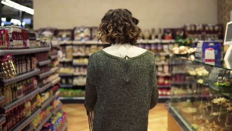 rare view of a woman with wavy hair is driving shopping trolley through food department in supermarket and looking around