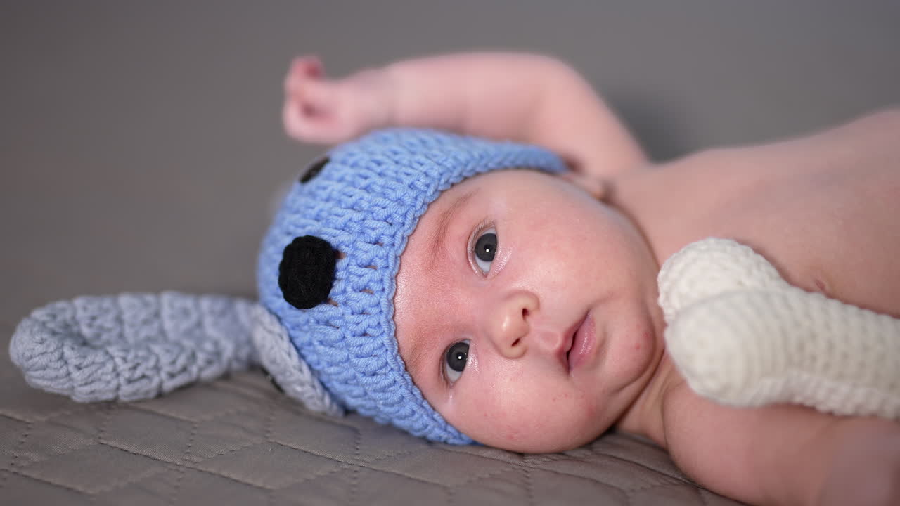 Small kid lying and moving his tiny hands quickly. Adorable babe in funny blue doggy cap and knitted bone. Beautiful child in the bedroom.
