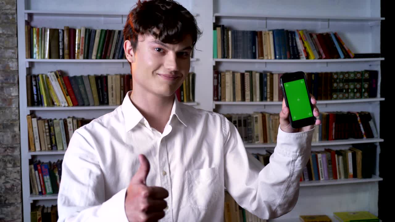 Brunette man is showing green screen of smartphone, like sign, watching at camera, communication concept, library on background