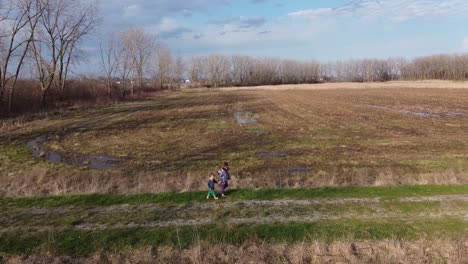 the kids walking in the wide grass field in the park of monroe michigan surrounded with withered trees under the bright cloudy sky - aerial shot