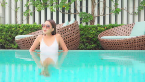 one pretty asian woman relaxing inside swimming pool leaning on her elbows at the pool's edge with luxury round rattan lounge chairs on the background in thailand hotel