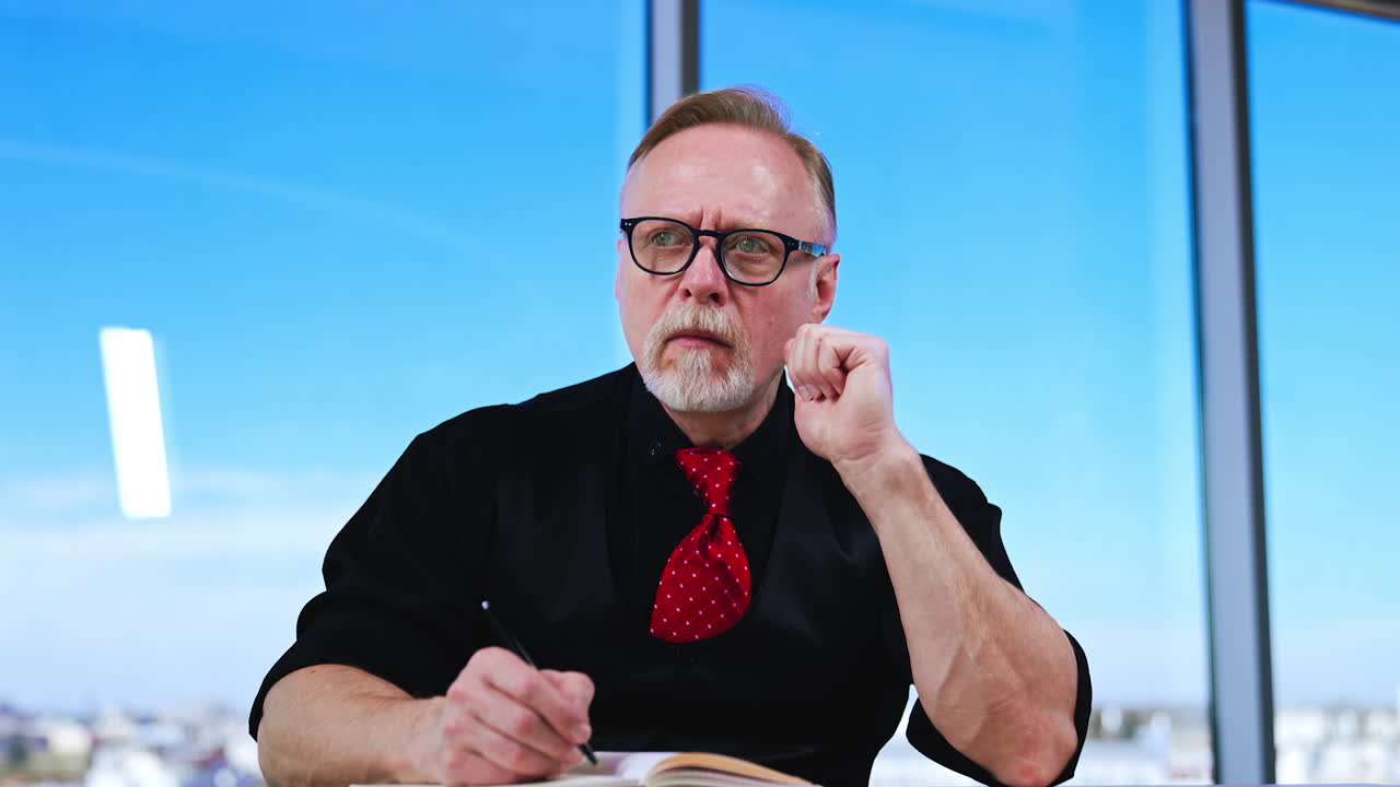 Thoughtful Caucasian mature man sits at desk. Businessman gets the idea and writes it down into the paper book.