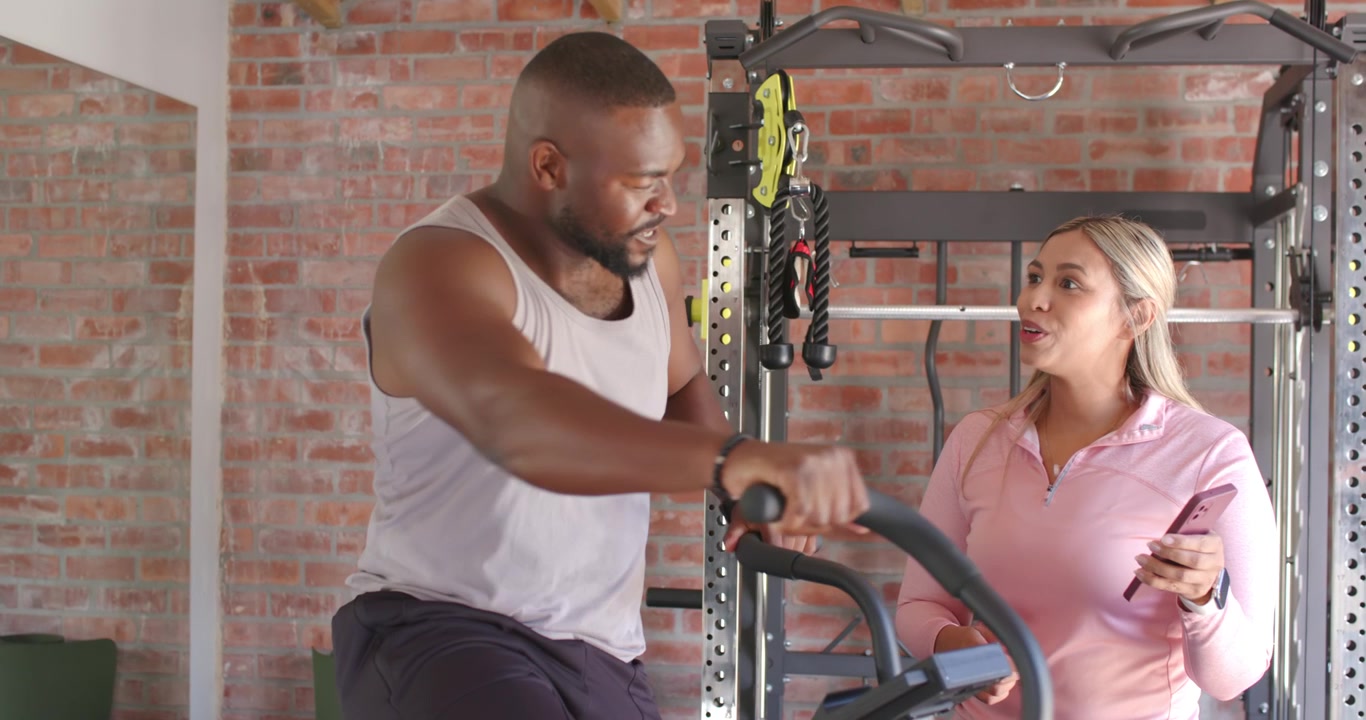 Diverse Partner trainieren im Home-Gym, Mann tritt Air Bike stärker, nachdem Frau Smartphone zeigt.