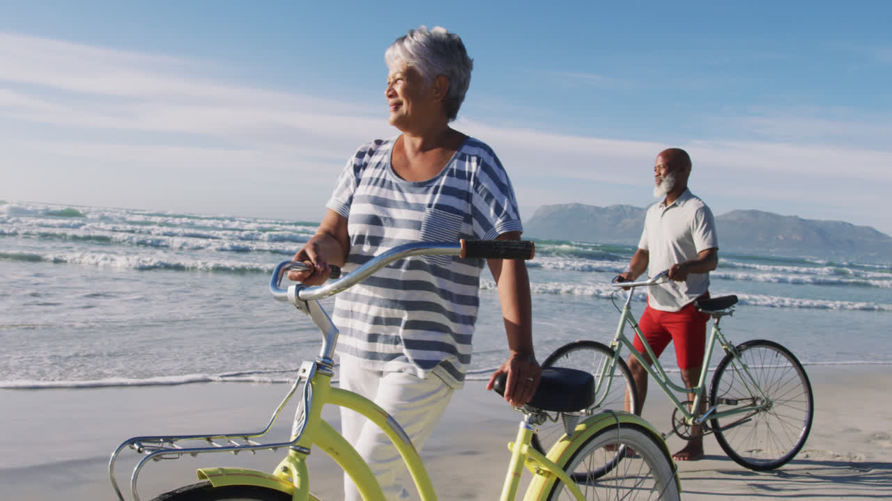 Smiling senior african american couple walking with bicycles at the beach