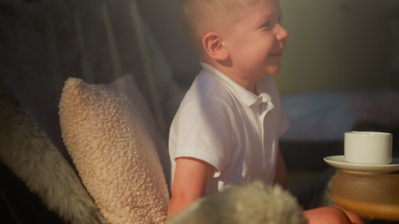 Child laughs sitting at table with cup. Joyful little boy rests in big comfortable chair in sunlit room. Smiling kid happy to spend morning at home