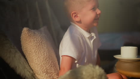 child laughs sitting at table with cup. joyful little boy rests in big comfortable chair in sunlit room. smiling kid happy to spend morning at home