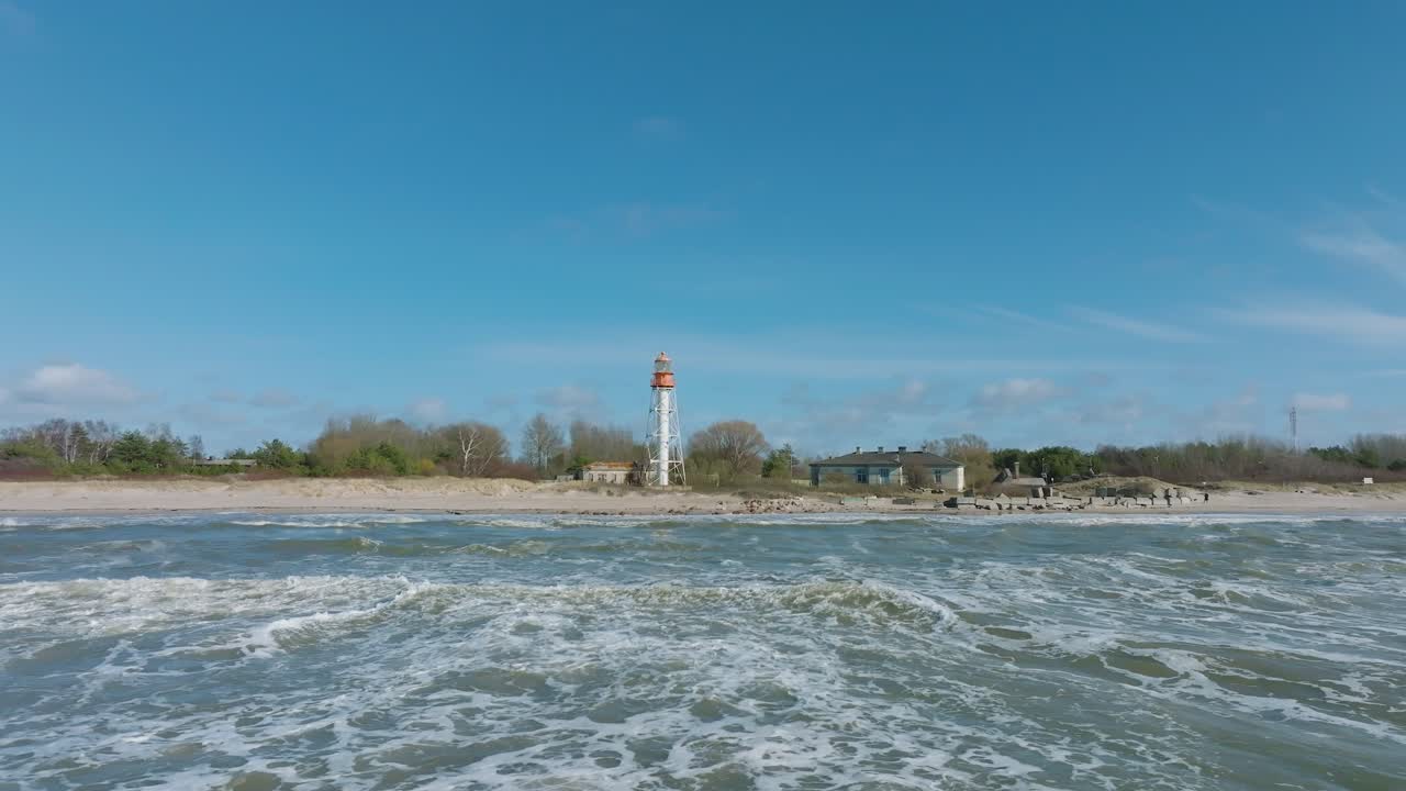 vista aérea del faro de color blanco, costa del mar báltico, letonia, playa de arena blanca, grandes olas chocando, día soleado con nubes, toma de órbita baja de drones
